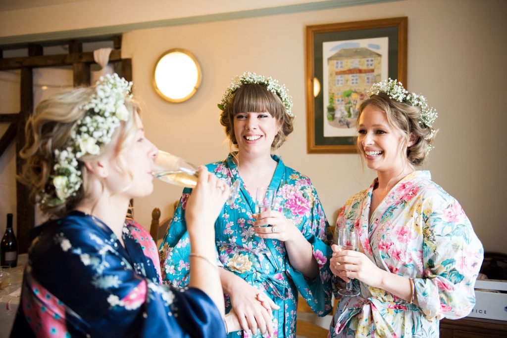 Bride with bridesmaids drink champagne wearing floral robes and floral crowns Driftwood Spars Wedding