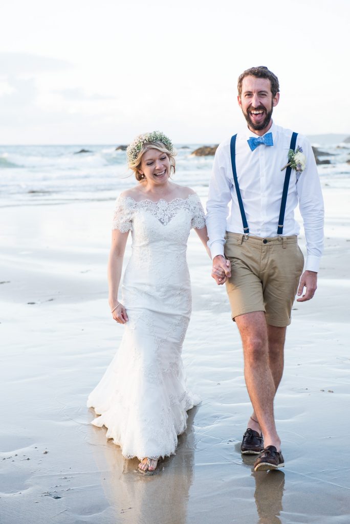 Happy boho bride with groom walking on Cornwall beach