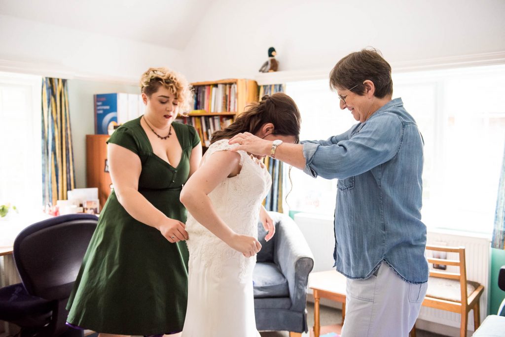 Mother and bridesmaid help bride into her white lace wedding dress