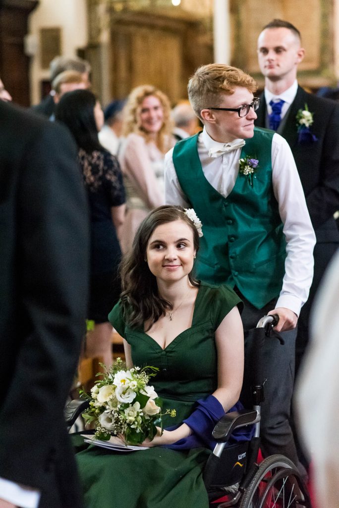 Bridal party follow the married couple. Groomsman escorts bridesmaid who sits in a wheel chair down the aisle.