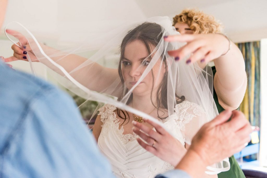 Bride in white lace dress is helped by her bridesmaid whilst putting on her veil