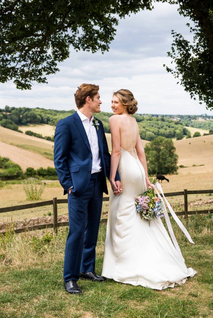 Surrey wedding couple laugh together. Bride wears a backless silk wedding dress and the groom is in a navy blue suit