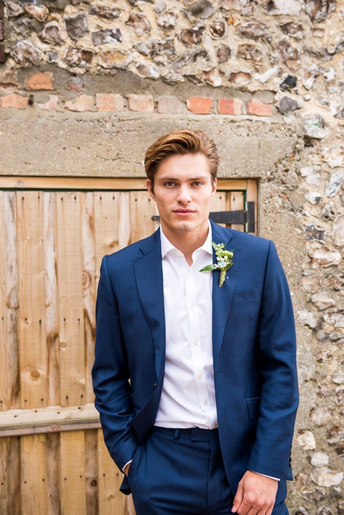 Groom stands in front of rustic barn dressed in a navy blue suit