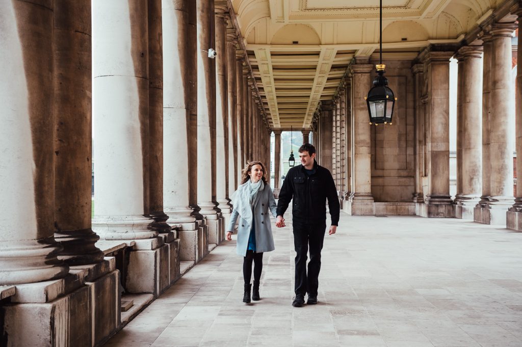 Engaged couple walk hand in hand down the colonnades of Greenwich Naval college 