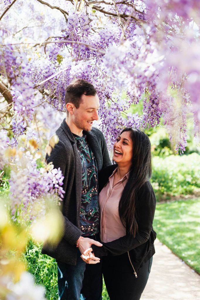 Engaged couple laugh together amongst purple wisteria flowers
