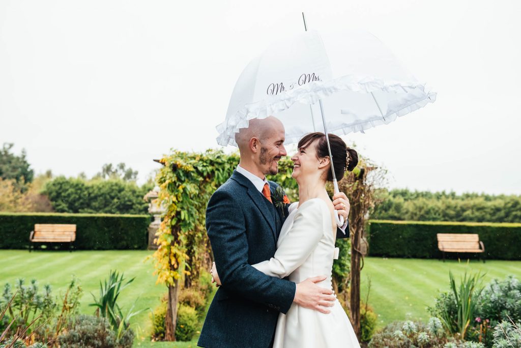 Couple embrace each other in the rain under an umbrella at their Surrey wedding