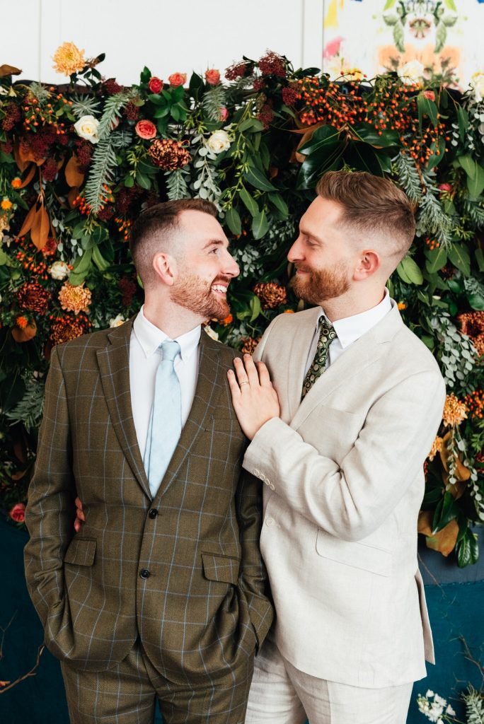 Grooms smile together in front of a flower wall backdrop