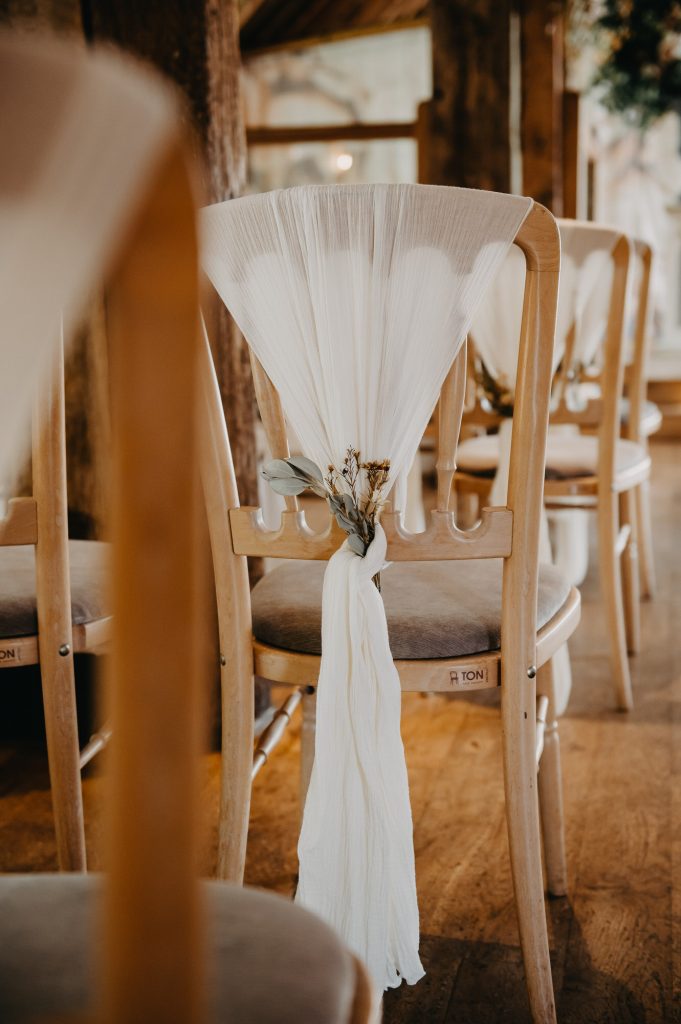 Modern wedding barn venue with light coloured wooden chairs and white floral backdrop. 