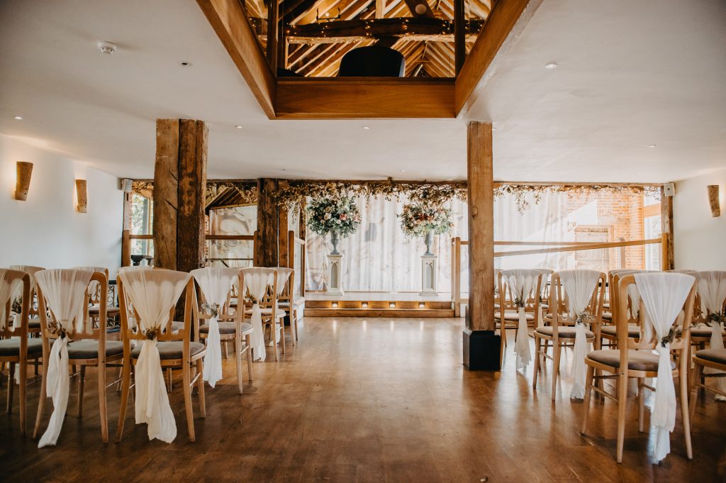 Modern wedding barn venue with light coloured wooden chairs and white floral backdrop. 