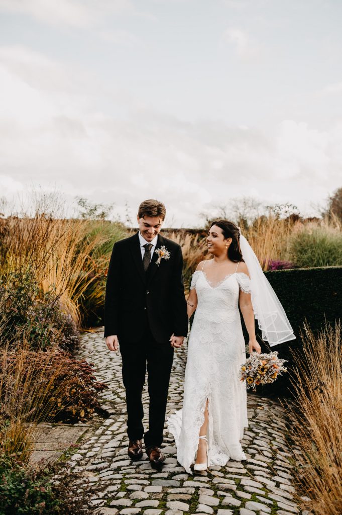 Couple walk hand in hand together through gardens of tall grasses and flowers.