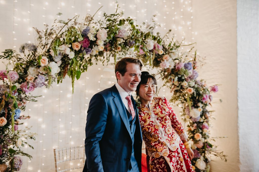 Couple Embrace With Floral Decor Behind - London Wedding Photography