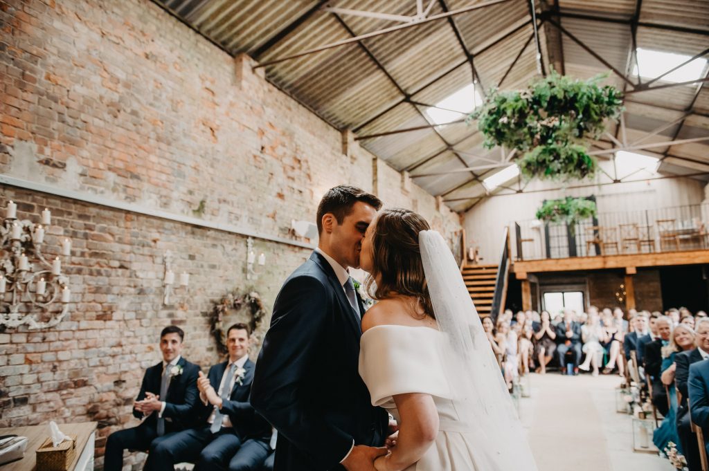 Couple exchange a first kiss in front of friends and family during relaxed wedding ceremony.