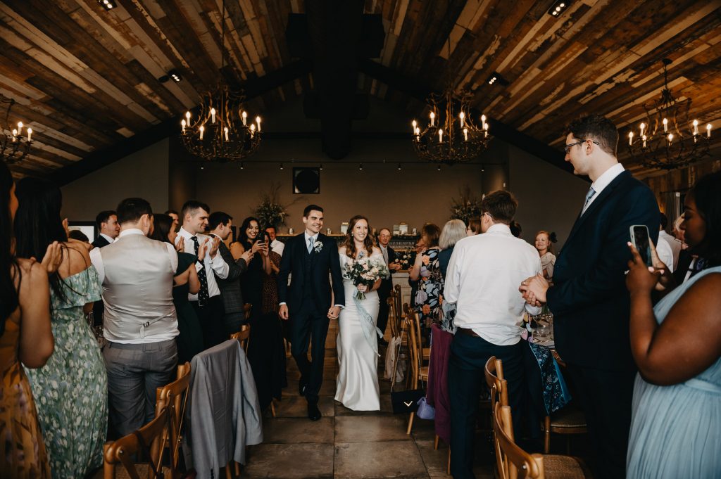 Couple enter their wedding breakfast room together walking down the table aisles. 