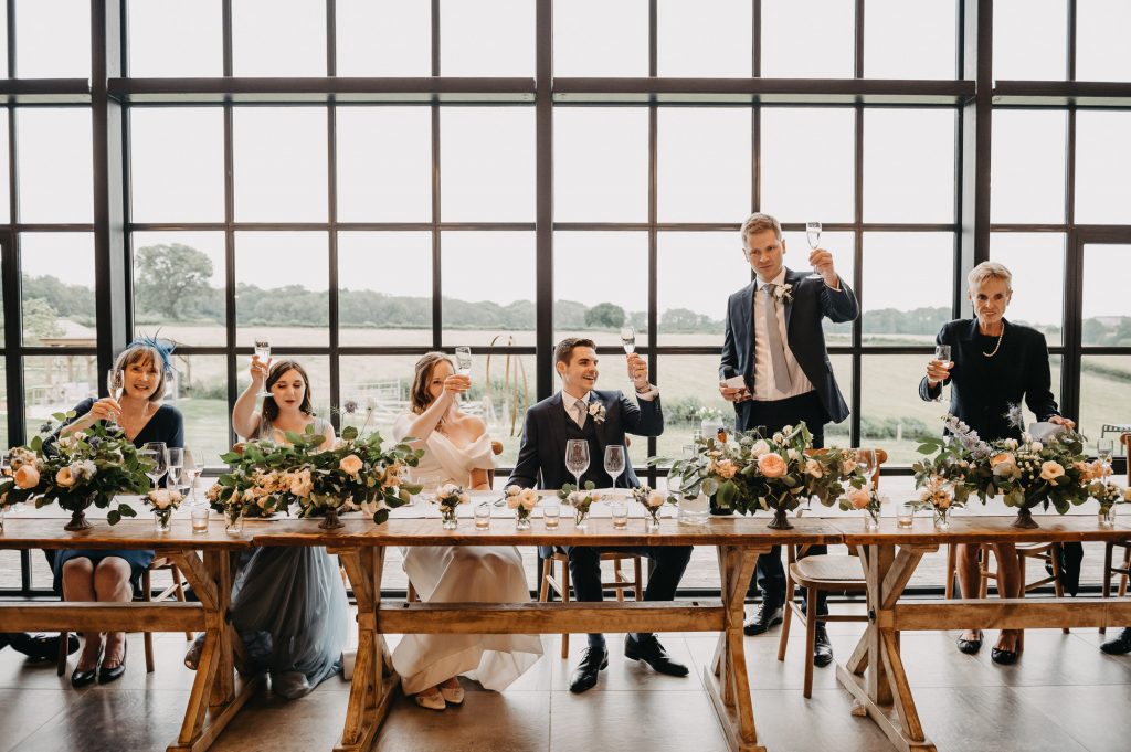 Best man stands up and gives a wedding speech from the top table. He is holding up his glass in a toast.