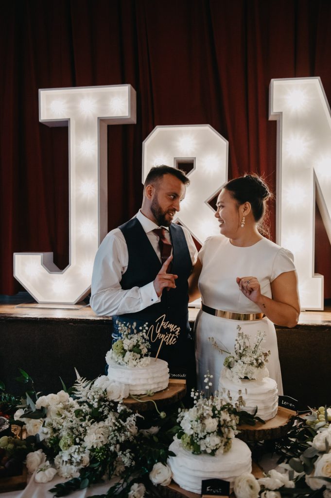 Couple Candidly Cut The Wedding Cake Together with Personalised Initial Signs Behind Them