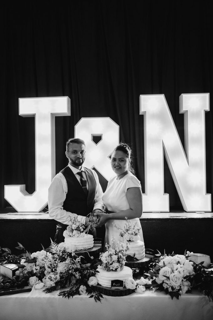 Couple Candidly Cut The Wedding Cake Together with Personalised Initial Signs Behind Them