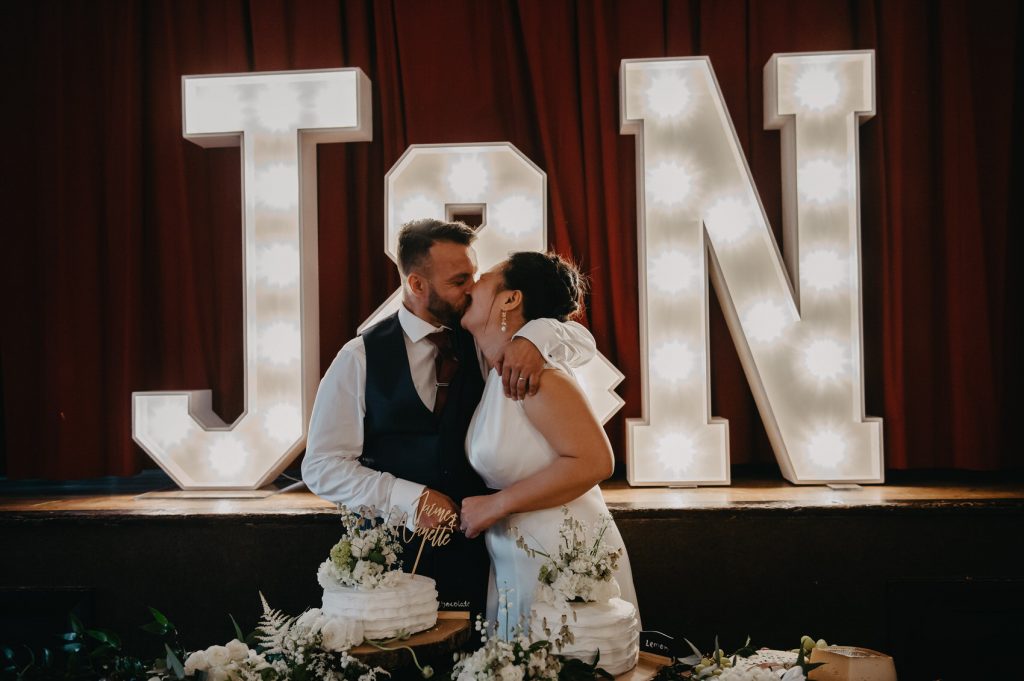 Couple Candidly Kiss Whilst They Cut The Wedding Cake Together with Personalised Initial Signs Behind Them